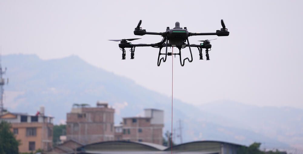 Drone hovering with a red line hanging below it, with buildings and hills in the background